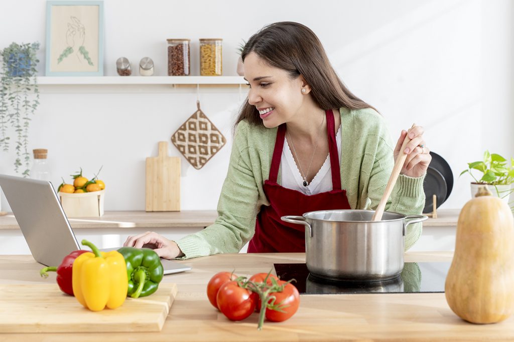 Aprendizaje en Youtube: ama de casa feliz removiendo una olla y leyendo una receta en un portátil rodeada de verduras frescas en una cocina moderna